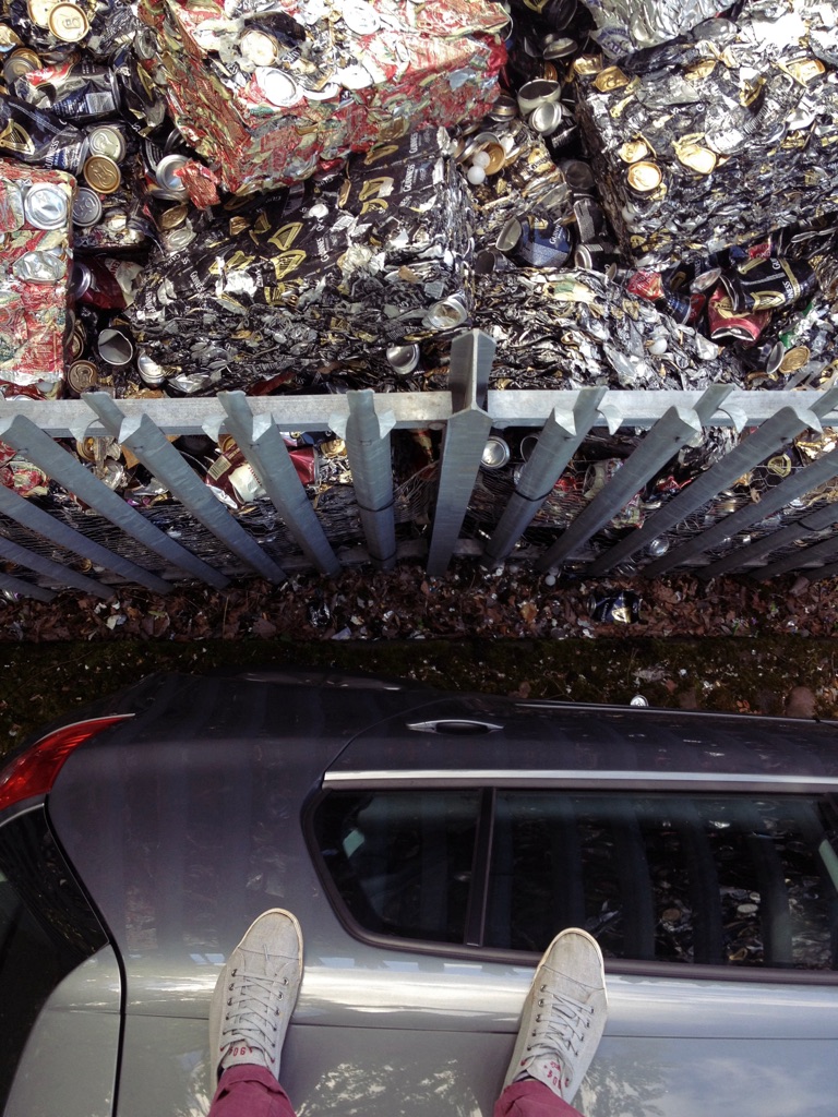 Photo taken with the camera pointing down revealing some cubes of crushed cans, and a metal fence separating the cans from a car, on which the photographer's stands